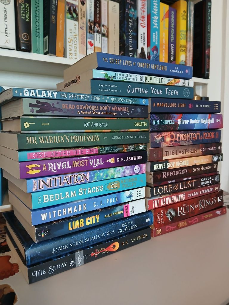 Two stacks of colorful books in front of a bookshelf, there is a small stack on top bridging the two stacks. Books shown are: The Secret Lives of Country Gentleman by KJ Charles. Burly Tales edited by Steve Berman. Cutting your Teeth by Caylan MacRae. Galaxy The Prettiest Star by Axelrod & Taylor. Dead Cowpokes Don't Wrangle edited by Mars Adler and H.S Wolfe. Kit and Basie by Tess Carletta. Mr Warren's Profession by Sebastian Nothwell. Love After The End edited by Joshua Whitehead. A Rival Most Vial by R.K. Ashwick. Initiation by Alethea Faust. Bedlam Stacks by Natasha Pulley. Witchmark by C.L. Polk. Liar City by Allie Therin. Dark Moon Shallow Sea by David R. Slayton. The Stray Spirit by R.K. Ashwick. A Marvelous Light by Freya Marske. Silver Under Nightfall by Rin Chupeco. Phantom and Rook by Aelina Isaacs. The Last Sun by KD Edwards. Brave Boy World edited by Lee Mandelo. Reforged by Seth Haddon. Finding Echoes by Foz Meadows. Lore and Lust by Karla Nikole. The Dawnhounds by Sascha Stronach. The Ruin of Kings by Jenn Lyons. Nimona by N.D Stevenson.