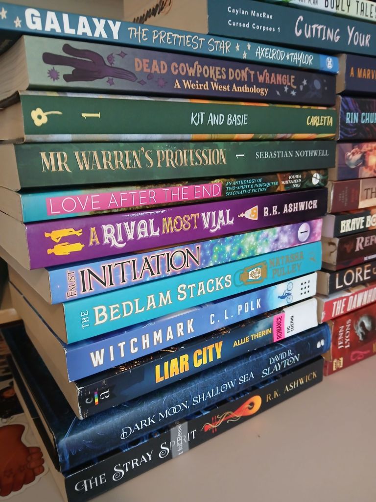 Two stacks of colorful books in front of a bookshelf, there is a small stack on top bridging the two stacks. Books shown in this closeup are: Galaxy The Prettiest Star by Axelrod & Taylor. Dead Cowpokes Don't Wrangle edited by Mars Adler and H.S Wolfe. Kit and Basie by Tess Carletta. Mr Warren's Profession by Sebastian Nothwell. Love After The End edited by Joshua Whitehead. A Rival Most Vial by R.K. Ashwick. Initiation by Alethea Faust. Bedlam Stacks by Natasha Pulley. Witchmark by C.L. Polk. Liar City by Allie Therin. Dark Moon Shallow Sea by David R. Slayton. The Stray Spirit by R.K. Ashwick. 