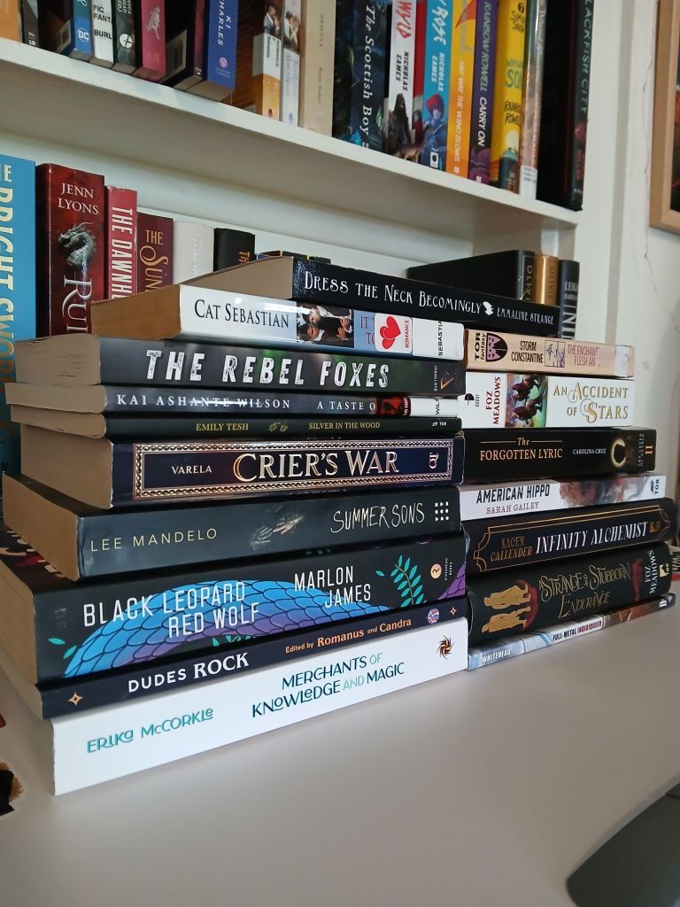 Two stacks of black and white books in front of a bookshelf. Top to bottom the books in left stack are: Dress the Neck Becomingly by Emmaline Strange. It takes two to tumble by Cat Sebastian. The Rebel Foxes by Noah Hawthorne. A Taste of Honey by Kai Ashante Wilson. Silver in the Wood by Emily Tesh. Crier's War by Nina Varela. Summer Sons by Lee Mandelo. Black Leopard Red Wolf by Marlon James. Dudes Rock edited by Romanus and Candra. Merchants of Knowledge and Magic by Erika McCorkle.
Top to bottom books on right stack are:  Dress the Neck Becomingly by Emmaline Strange. The Enchaments of Flesh and Stone by Storm Constantine. The Forgotten Lyric by Carolina Cruz. American Hippo by Sarah Gailey. Infinity Alchemist by Kacen Callender. A Strange and Stubborn Endurance by Foz Meadows. Full Metal Indigiqueer by Joshua Whitehead.