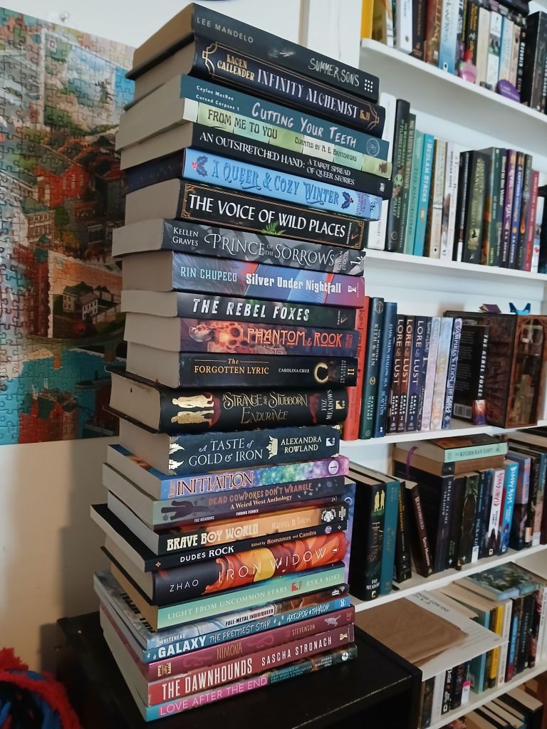 A stack of books beside a bookshelf with a puzzle on the wall. Top to bottom books are:
Summer Sons by Lee Mandelo.
Infinity Alchemist by Kacen Callender.
Cutting Your Teeth by Caylan McRae.
From Me to You edited by A.E. Bratchford.
An Outstretched Hand curated by Write Here Right Queer.
A Queer and Cozy Winter curated by Rainbow Crate.
The Voice of Wild Places by Noah Hawthorne.
Prince of Sorrows by Kellen Graves.
Silver Under Nightfall by Rin Chupeco.
The Rebel Foxes by Noah Hawthorne. Phantoms and Rook by Aelina Isaacs.
The Forgotten Lyric by Carolina Cruz.
A Strange and Stubborn Endurance by Foz Meadows.
A Taste of gold and iron by Alexandra  Rowland.
Initiation by Alethea Faust.
Dead Cowpokes Don't Wrangle curated by Mars Adler and H.s. Wolfe.
Finding Echoes by Foz Meadows.
Brave Boy World edited by Lee Mandelo.
Dudes Rock curated by Romanus and Candra.
Iron Widow by Xiran Zhao.
Light From Uncommon Stars by Ryka Aoki.
Full Metal Indigiqueer by Joshua Whitehead.
Galaxy by Jadzia Axelrod.
Nimona by N.D. Stevenson.
The Dawnhounds by Sascha Stronach.
Love After the end by Joshua Whitehead.