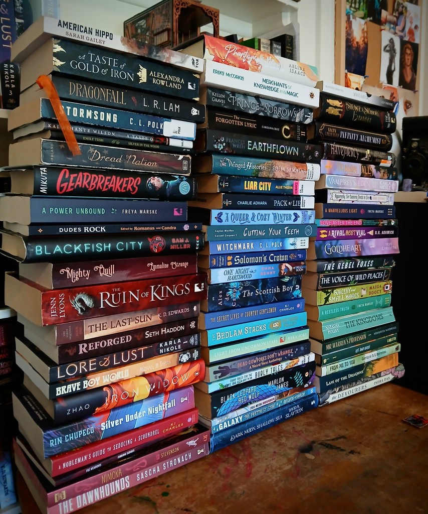 Three stacks of books in front of a bookshelf. Left shelf, top down. American Hippo by Sarah Gailey. A Taste of Gold and Iron by Alexandra Rowland. Dragonfall by L.R. Lam. Stormsong by C.L. Polk. A Taste of Honey by Kai Ashante Wilson. Silver in the Wood by Emily Tesh. Dread Nation by Justina Ireland. Gearbreakers by Zoe Hana Mikuta. A Power Unbound by Freya Marske. Dudes Rock edited by Romanus. Blackfish by Sam J. Miller. Mighty Quill by Emmaline Strange. The Ruin of Kings by Jenn Lyons. The Last Sun by K.D. Edwards. Reforged by Seth Haddon. Lore and Lust by Karla Nikole. Brave Boy World. Iron Widow by Xiran Zhao. Silver Under Nightfall by Rin Chupeco. A Nobleman's Guide to Seducing a Scoundrel by KJ Charles. Finding Echoes by Foz Meadows. Nimona by ND Stevenson. The Dawnhounds by Sascha Stronach. Middle Stack, top down. Phantom and Rook by Aelina Isaacs. Merchants of Knowledge and Magic by Erika McCorkle. Prince of Sorrows by Kellen Graves. The Forgotten Lyric by Carolina Cruz. Earthflown by Frances Wren. The Winged Histories Sofia Samatar. Liar City by Allie Therin. Pack of Lies by Charlie Adhara. A Queer and Cozy Winter curated by Rainbow Crate. Cutting Your Teeth by Caylan Macrae. Witchmark by C.L. Polk. Solomon's Crown by Natasha Siegel. The Scottish Boy by Alex de Campi. The Secret Lives of Country Gentlemen by K.J. Charles. Light from Uncommon Stars by Ryka Aoki. Dead Cowpokes Don't Wrangle curated by H.S. Wolfe and Mars Adler. Burly Tales edited by Steve Berman. Full Metal Indigiqueer edited by Joshua Whitehead. Galaxy The Prettiest Star by Jadzia Axelrod. Dark Moon Shallow Sea by David Slayton. Soul Eater by Lily Mayne. A Strange and Stubborn Endurance by Foz Meadows. Infinity Alchemist by Kacen Callendar. Havemercy by Jadzia Jones. Dragon Falls by Davis Lavender. Vampires of Eden by Karla Nikole. Apparently, Sir Cameron must Die by Greer Stothers. A Marvellous Light by Freya Marske. Love After the End edited by Joshua Whitehead. A Rival Most Vial by R.K. Ashwick. Goldheart by Tess Carletta. The Rebel Foxes by Noah Hawthorne. The Voice of Wild Places by Noah Hawthorne. Sorcery and Small Magics by Maiga Doocy. A Restless Truth by Freya Marske. The Kingdoms by Natasha Pulley. Mr Warren's Profession by Sebastian Nothwell. Kit and Basie by Tess Carletta. From Me to You an Anthology of Human Connections. Law of the Dragons by Y.W. Lumaris. The Devourers by Indra Das.