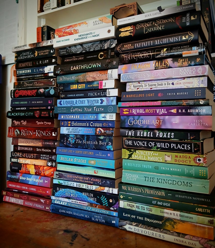 Three stacks of books in front of a bookshelf. Left shelf, top down. American Hippo by Sarah Gailey. A Taste of Gold and Iron by Alexandra Rowland. Dragonfall by L.R. Lam. Stormsong by C.L. Polk. A Taste of Honey by Kai Ashante Wilson. Silver in the Wood by Emily Tesh. Dread Nation by Justina Ireland. Gearbreakers by Zoe Hana Mikuta. A Power Unbound by Freya Marske. Dudes Rock edited by Romanus. Blackfish by Sam J. Miller. Mighty Quill by Emmaline Strange. The Ruin of Kings by Jenn Lyons. The Last Sun by K.D. Edwards. Reforged by Seth Haddon. Lore and Lust by Karla Nikole. Brave Boy World. Iron Widow by Xiran Zhao. Silver Under Nightfall by Rin Chupeco. A Nobleman's Guide to Seducing a Scoundrel by KJ Charles. Finding Echoes by Foz Meadows. Nimona by ND Stevenson. The Dawnhounds by Sascha Stronach. Middle Stack, top down. Phantom and Rook by Aelina Isaacs. Merchants of Knowledge and Magic by Erika McCorkle. Prince of Sorrows by Kellen Graves. The Forgotten Lyric by Carolina Cruz. Earthflown by Frances Wren. The Winged Histories Sofia Samatar. Liar City by Allie Therin. Pack of Lies by Charlie Adhara. A Queer and Cozy Winter curated by Rainbow Crate. Cutting Your Teeth by Caylan Macrae. Witchmark by C.L. Polk. Solomon's Crown by Natasha Siegel. The Scottish Boy by Alex de Campi. The Secret Lives of Country Gentlemen by K.J. Charles. Light from Uncommon Stars by Ryka Aoki. Dead Cowpokes Don't Wrangle curated by H.S. Wolfe and Mars Adler. Burly Tales edited by Steve Berman. Full Metal Indigiqueer edited by Joshua Whitehead. Galaxy The Prettiest Star by Jadzia Axelrod. Dark Moon Shallow Sea by David Slayton. Soul Eater by Lily Mayne. A Strange and Stubborn Endurance by Foz Meadows. Infinity Alchemist by Kacen Callendar. Havemercy by Jadzia Jones. Dragon Falls by Davis Lavender. Vampires of Eden by Karla Nikole. Apparently, Sir Cameron must Die by Greer Stothers. A Marvellous Light by Freya Marske. Love After the End edited by Joshua Whitehead. A Rival Most Vial by R.K. Ashwick. Goldheart by Tess Carletta. The Rebel Foxes by Noah Hawthorne. The Voice of Wild Places by Noah Hawthorne. Sorcery and Small Magics by Maiga Doocy. A Restless Truth by Freya Marske. The Kingdoms by Natasha Pulley. Mr Warren's Profession by Sebastian Nothwell. Kit and Basie by Tess Carletta. From Me to You an Anthology of Human Connections. Law of the Dragons by Y.W. Lumaris. The Devourers by Indra Das.